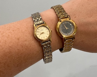May include: Two vintage wristwatches. One watch has a gold-tone case and band with a cream-colored face. The other watch has a gold-tone case and band with a dark gray face. Both watches are on a wrist.