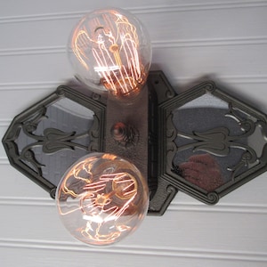 May include: A vintage-style wall sconce with two exposed filament light bulbs and a decorative mirrored backplate. The sconce is made of dark metal and has a geometric design.