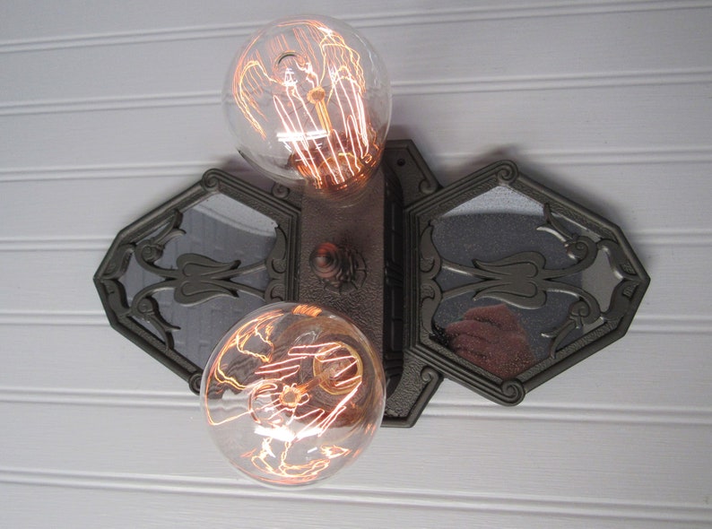 May include: A vintage-style wall sconce with two exposed filament light bulbs and a decorative mirrored backplate. The sconce is made of dark metal and has a geometric design.