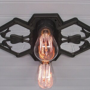 May include: A black metal ceiling light fixture with an ornate design. The fixture has two exposed light bulbs with a filament design.