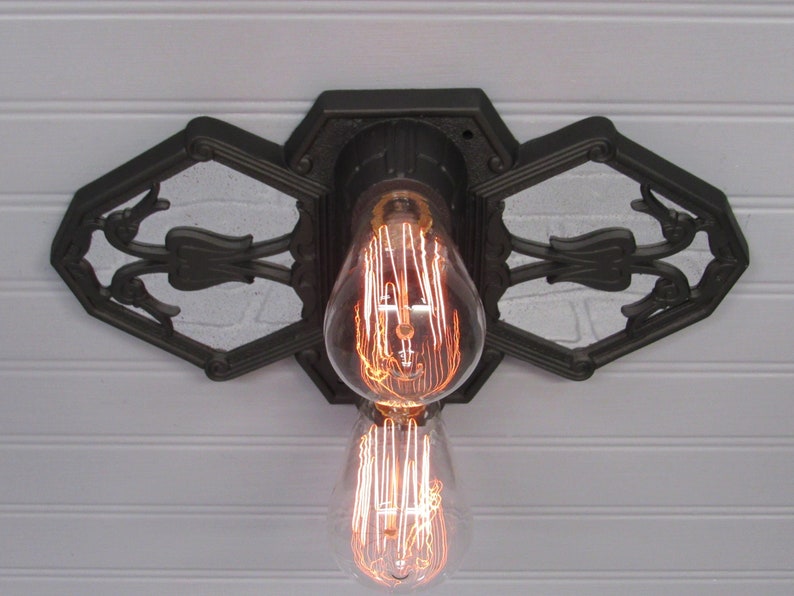 May include: A black metal ceiling light fixture with an ornate design. The fixture has two exposed light bulbs with a filament design.