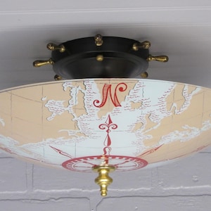 Puede incluir: Una lámpara de techo vintage con temática náutica. La pantalla de cristal presenta un diseño de mapa, una rosa de los vientos y la letra "N". La lámpara tiene una base negra con detalles en latón y un diseño de timón. Ideal para un ambiente marítimo.