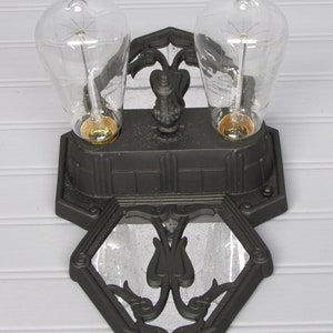 May include: A black metal wall sconce with two clear glass light bulbs. The sconce has a decorative design with a mirrored center.