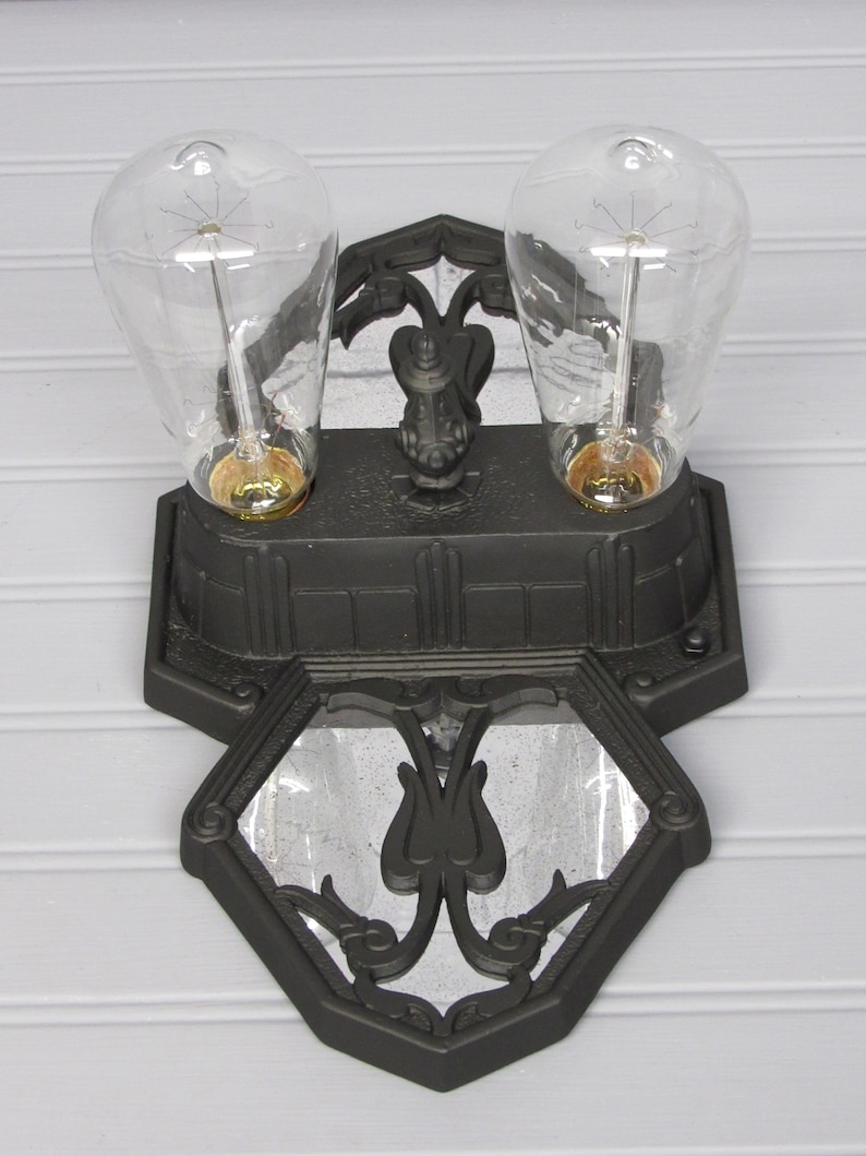May include: A black metal wall sconce with two clear glass light bulbs. The sconce has a decorative design with a mirrored center.