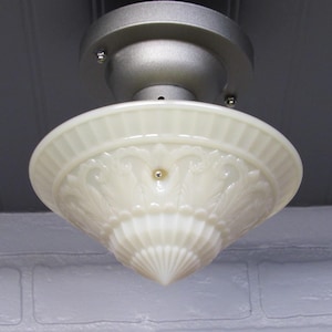 May include: Cream-colored, vintage-style ceiling light fixture. The glass shade has a decorative, embossed pattern and a ribbed edge. The fixture is mounted on a silver-colored metal base. The light is hanging from a gray ceiling.