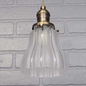 May include: A frosted glass pendant light shade with a ribbed design hangs from a brass ceiling fixture. The shade is white and has a bell shape.
