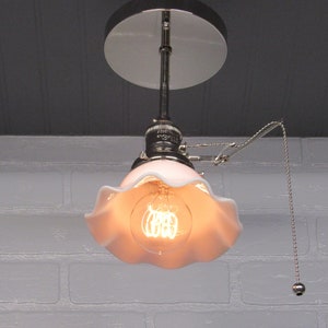 May include: A white ceiling light fixture with a pull chain switch and a white ruffled glass shade. The light is on and the bulb is visible.