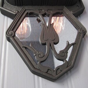 May include: A black metal wall sconce with a geometric glass panel featuring a stylized floral design. The sconce has two visible light bulbs.