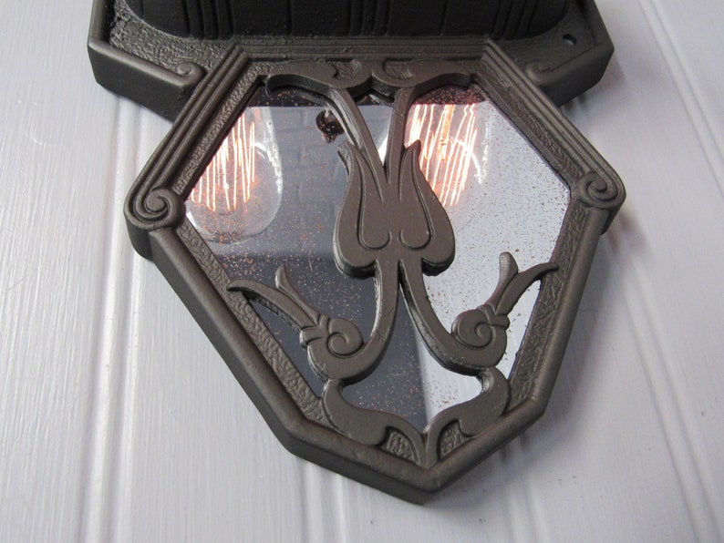 May include: A black metal wall sconce with a geometric glass panel featuring a stylized floral design. The sconce has two visible light bulbs.