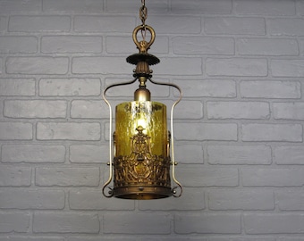 May include: A hanging pendant light fitting featuring a cylindrical amber-coloured textured glass shade encased in an ornate bronze-toned metal frame with intricate scrollwork details, suspended by a matching metal chain and canopy.