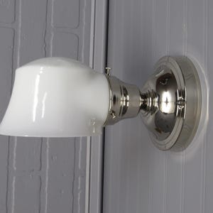 May include: A wall-mounted light fixture with a white, bell-shaped glass shade and a polished nickel finish. The fixture is mounted on a round, decorative base. The light is designed for indoor use.