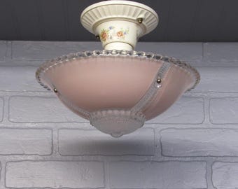 May include: A semi-flush mount ceiling light fixture featuring a pale pink glass bowl with a clear, textured glass rim and a central finial. The cream-colored metal canopy is decorated with a delicate floral pattern of small roses and leaves.