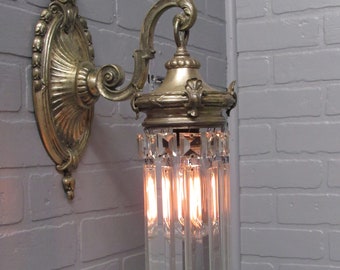 May include: A vintage brass wall sconce with a crystal chandelier-style shade. The sconce has a decorative scroll design and features a single light bulb.