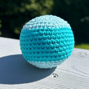 May include: A crocheted ball in shades of light blue and turquoise. The handmade ball has a striped design, with a band of turquoise in the middle. The ball is sitting on a white surface, with a blurred green background.