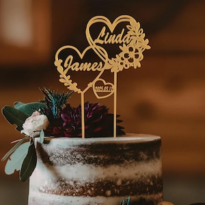 May include: Gold wedding cake topper featuring two intertwined hearts. The hearts are adorned with floral designs and the names "Linda" and "James" are inscribed on them. The date "2023.03.12" is also inscribed on the topper.