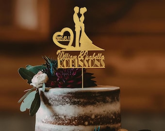 May include: A gold-colored wedding cake topper featuring a silhouette of a couple, a heart with the date 2025.03.11, and the names William & Isabella ROBINSONS. The topper is placed on a decorated cake with flowers.