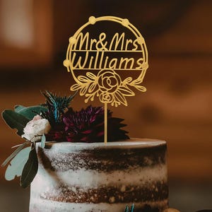 Puede incluir: Topper de pastel de bodas dorado con un dise&ntilde;o circular y el texto "Mr. & Mrs. Williams" y detalles florales.