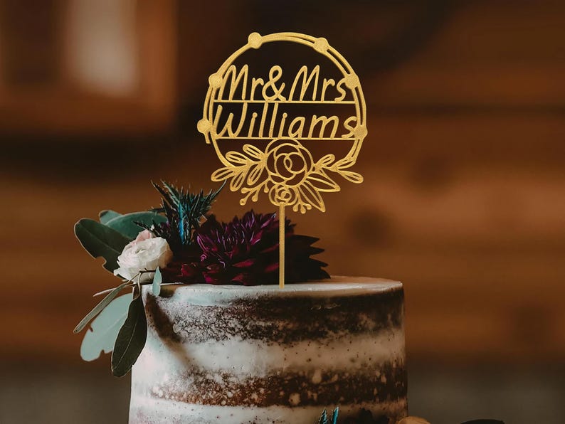 Puede incluir: Topper de pastel de bodas dorado con un dise&ntilde;o circular y el texto "Mr. & Mrs. Williams" y detalles florales.