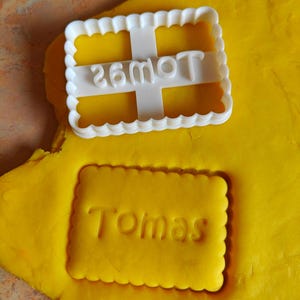 May include: A white plastic cookie cutter with a scalloped edge and a cross design, with the text "26moT". Below, a yellow dough has been cut into a rectangular shape with the name "Tomas".