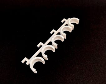 May include: White plastic pipe holder with six slots for organizing pipes. The holder is designed to keep pipes organized and prevent them from rolling around.