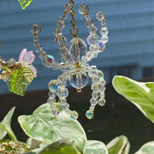 May include: A crystal spider hanging from a gold chain. The spider is made of clear crystals with iridescent accents. The spider is hanging from a gold chain and is surrounded by green leaves.