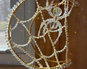 May include: A handcrafted suncatcher with a gold-coloured crescent moon and spider web design. The moon and web are adorned with clear, faceted beads. A large, clear bead spider is suspended in the centre. The suncatcher hangs from a delicate chain.