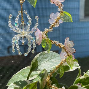 May include: A delicate crystal spider hanging from a beaded and wired floral garland with pink and green glass flowers and leaves.