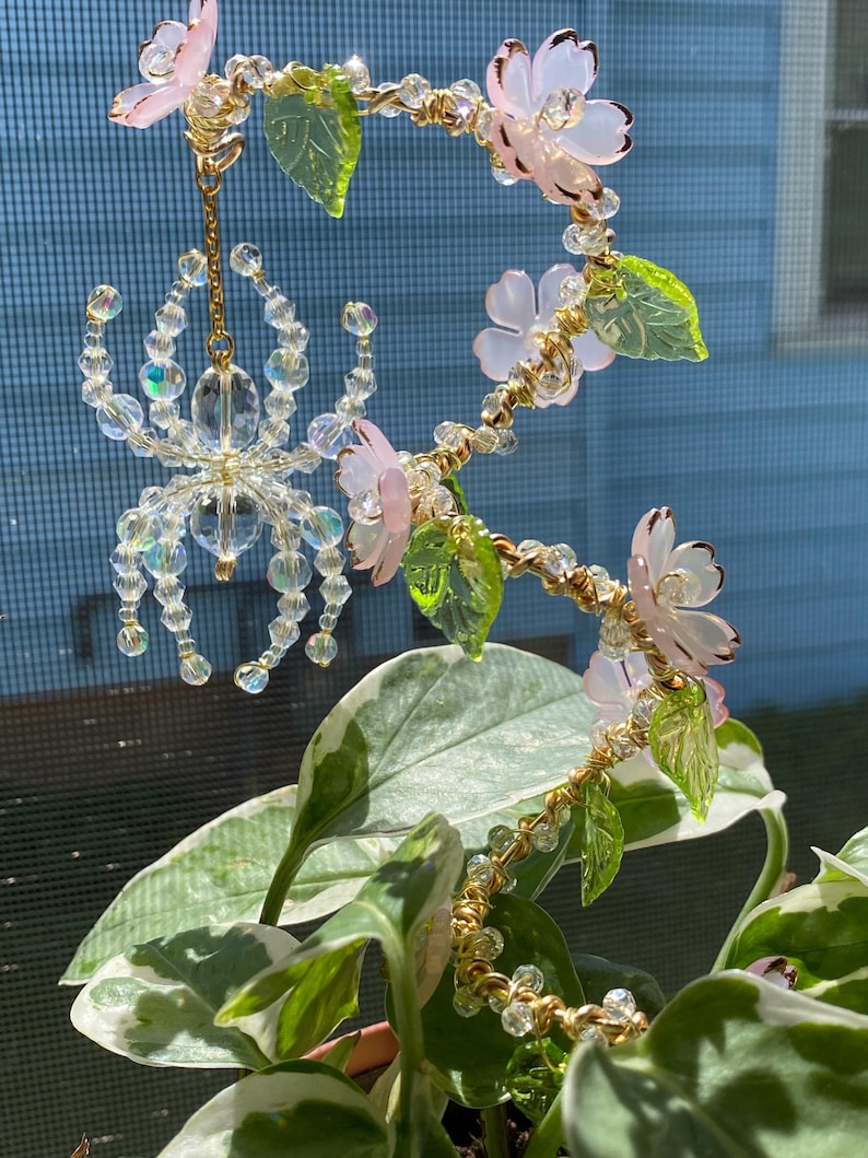May include: A delicate crystal spider hanging from a beaded and wired floral garland with pink and green glass flowers and leaves.