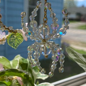 May include: A crystal spider ornament with a gold chain hanging from a plant. The spider is made of clear iridescent beads and has a gold wire frame.
