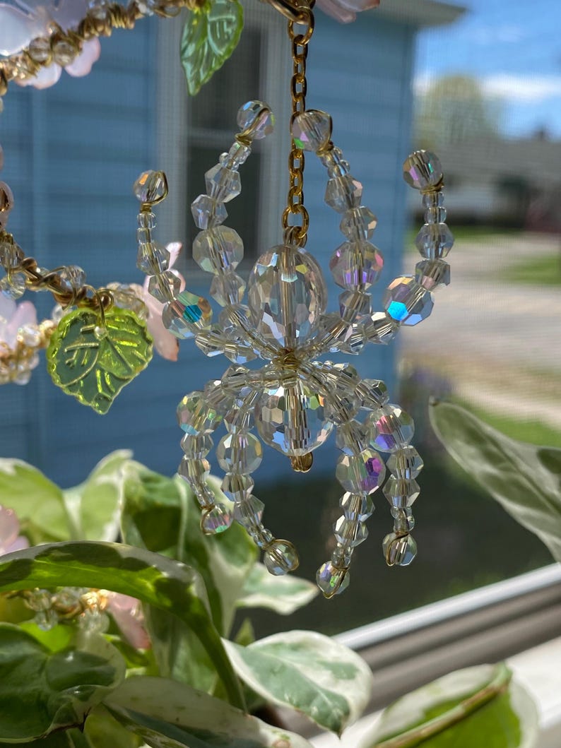 May include: A crystal spider ornament with a gold chain hanging from a plant. The spider is made of clear iridescent beads and has a gold wire frame.