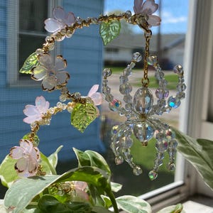 May include: A crystal spider hanging from a beaded floral vine with pink and green accents. The vine is made of wire and beads, and the spider is made of clear crystals.