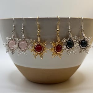 以下が含まれることがあります： 手作りのイヤリングのコレクション。それぞれ太陽のモチーフが特徴です。ローズクォーツ、赤いカーネリアン、緑色の石など、さまざまな宝石が銀色と金色のワイヤーでセットされています。イヤリングは陶器のボウルに飾られています。