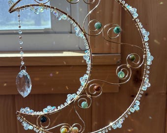 May include: A crescent moon-shaped wall hanging crafted from gold-colored wire. The moon is adorned with clear, iridescent beads and colorful gemstones. A clear crystal hangs from a chain at the bottom. The suncatcher is hanging in front of a window.