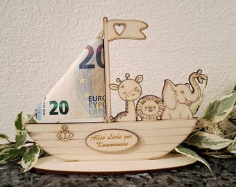 Könnte beinhalten: Ein hölzerner Bootsförmiger Geldhalter mit einem Segel aus einem Zwanzig-Euro-Schein. Das Boot zeigt ausgeschnittene Tierfiguren: eine Giraffe, ein Löwe und ein Elefant. Ein Banner mit einem Herzen und dem Text "Alles Liebe zur Kommunion" ist ebenfalls vorhanden.