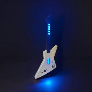 May include: A white electric guitar with a unique, angular design. The guitar features blue LED lights along the neck and body, creating a futuristic aesthetic. The instrument is set against a dark background, with a blue glow emanating from the base.