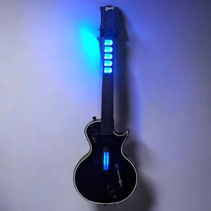 May include: A black electric guitar with a white trim, illuminated by blue LED lights. The guitar is mounted on a gray wall, with the Gibson logo visible on the headstock. The lights create a striking visual effect.