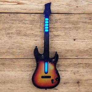May include: A toy electric guitar with a sunburst finish, black accents, and a blue glowing neck and body. The guitar is set against a wooden background.