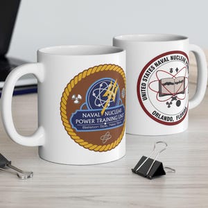 Puede incluir: Dos tazas de cerámica blanca con emblemas azul marino y dorados. Una taza presenta el texto "NAVAL NUCLEAR POWER TRAINING UNIT" y la otra dice "UNITED STATES NAVAL NUCLEAR" con "ORLANDO, FLORIDA".