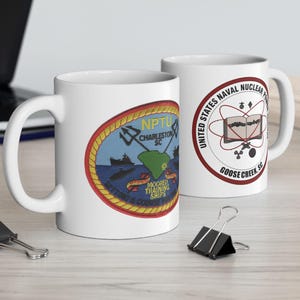 Puede incluir: Dos tazas de cerámica blanca. Una taza presenta un emblema colorido con el texto "NPTU Charleston SC" y "Moored Training Ships". La otra taza tiene un diseño circular con "United States Naval Nuclear Power" y "Goose Creek SC".