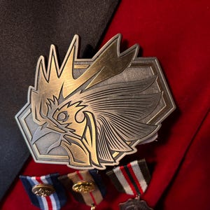 May include: A gold and silver metal pin featuring a stylized design of a bird's head with a crest. The pin is attached to a red fabric background.