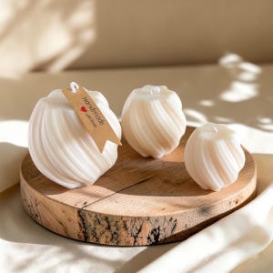 Könnte beinhalten: Drei cremeweiße, kugelförmige Kerzen mit einer vertikalen, spiralförmigen Rippenstruktur. Die größte Kerze hat ein braunes, rechteckiges Papieretikett mit dem Text "Handmade with love" und einem kleinen roten Herzsymbol.