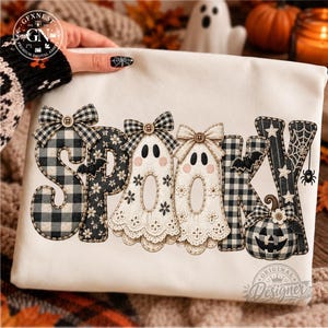 May include: A white sweatshirt featuring the word SPOOKY in a decorative appliqué style. The letters are designed with black and white buffalo plaid, floral patterns, and two ghost shapes. The design includes bows, a pumpkin, bats, and a spider web.