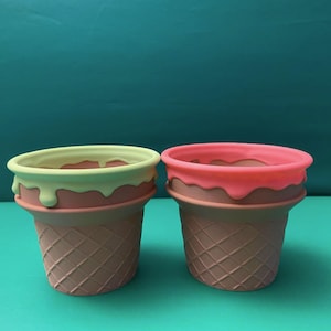 May include: Two silicone ice cream cone shaped cups. One cup has a light green rim and the other has a pink rim.
