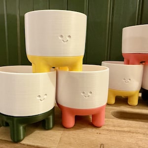 May include: Five white ceramic planters with a smiley face design. Each planter has three legs and is painted in a different colour: yellow, red, green, yellow, and black.
