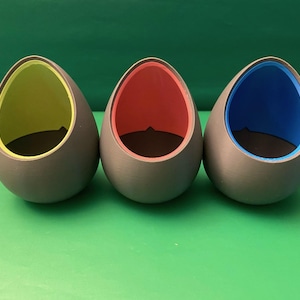May include: Three grey ceramic egg-shaped planters with a smooth finish. The planters have a coloured interior, one yellow, one red, and one blue.