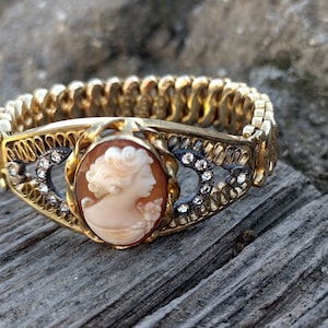 Può includere: Un bracciale cammeo dorato con un profilo scolpito di donna. Il cammeo ovale è incastonato in una cornice decorativa con cristalli trasparenti. Il bracciale ha un design flessibile e collegato.