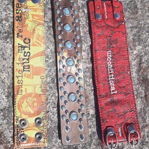 May include: Three leather bracelets. The first bracelet is yellow and orange with a print of a motorcycle and the text "Let go of my music it's my release". The second bracelet is brown with turquoise stones and silver studs. The third bracelet is red with a textured surface and the text "Unconditional".