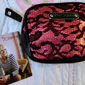 May include: A pink and black lace cosmetic bag with a black zipper and a silver metal tag that reads "Betsey Johnson".