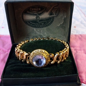 May include: A gold-toned adjustable bracelet with a central light blue stone, displayed in a vintage box. The box is dark green with the text "CARMEN ADJUSTABLE BRACELET". The bracelet has a detailed, ornate design.