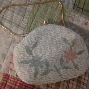 Op de afbeelding: Een vintage, crèmekleurige, met kralen bezette handtas met een gouden ketting. De tas heeft bloemenborduurwerk in blauw, roze en groen. De sluiting is goudkleurig. Een retro accessoire uit de jaren 50-60.